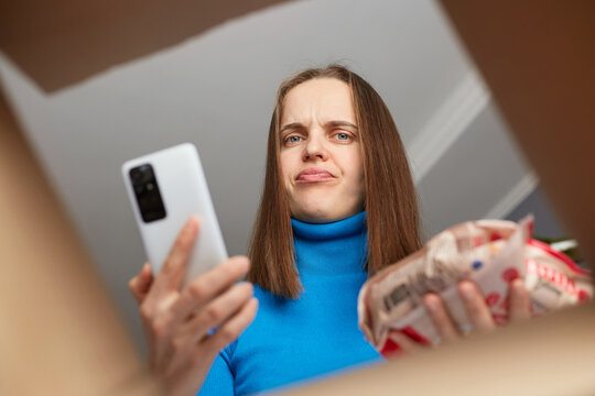 Online home delivery, internet orders and grocery shopping. Disappointed frustrated woman customer receive wrong order of groceries, looking upset inside box and pouting, holding cell phone in hands. - Powered by Adobe