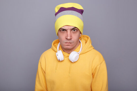 Image of angry aggressive Caucasian hipster man wearing beanie hat and yellow casual hoodie, arguing with somebody, looking at camera with clenched teeth, standing isolated over gray background.