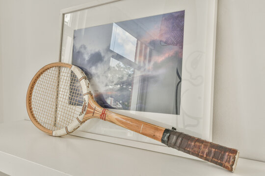 Amsterdam, Netherlands - 10 April, 2021: an old tennis racket on a white shelf next to a framed photograph of a cloudy sky in the background