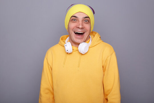Indoor shot of extremely happy joyful man wearing beanie hat and yellow casual hoodie standing isolated over gray background, looking at camera, laughing, being in good mood, expressing happiness.