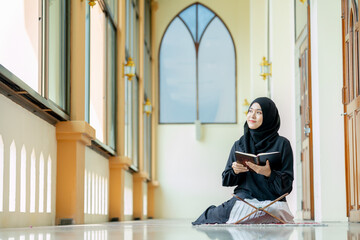 The image of an Asian Muslim woman in the Islamic religion in hijab in black color. Sitting reading the Quran and having a happy smiling face Staying in a beautiful mosque out of respect for God.