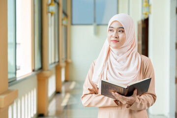 The image of an Asian Muslim woman in the Islamic religion in hijab in cream color. Standing reading the Qur'an and having a happy smiling face Staying in a beautiful mosque out of respect for God.