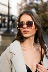 close-up portrait of a charming brunette on the street in spring, wearing a stylish beige coat, a top with razors and sunglasses
