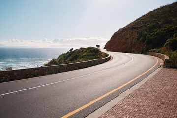 Fototapeta premium Mountain, road and ocean view with no people for travel, destination or sightseeing in Cape Town. Nature, beauty on empty street for road trip, vacation or holiday on South Africa blue sky background
