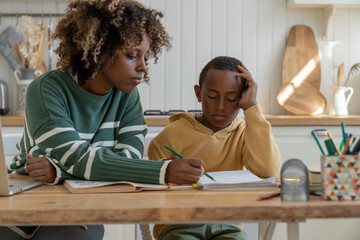 Focused biracial mother explaining difficult school task to attentive son for effective learning. African American personal tutor woman and little schoolboy sit at desk in home kitchen study together