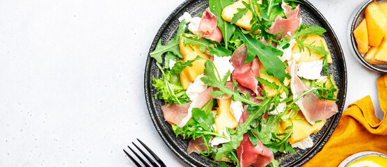Delicious melon salad with cantaloupe, prosciutto, soft cheese and arugula on white table...