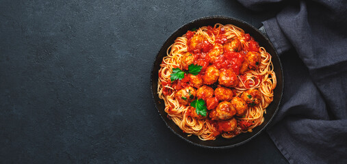Spaghetti pasta with meatballs in tomato sauce with parsley in frying pan, dark table background, top view. Banner, copy space