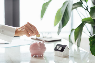 Hand putting money in White piggy bank with tiny house on white marble background and blurred bokeh...