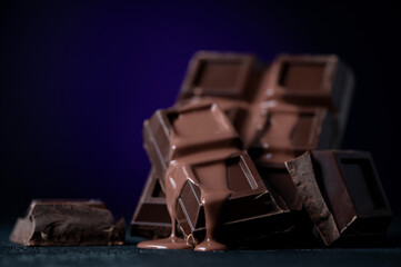 Broken chocolate bar is poured with melted chocolate. Chocolate sauce. Belgian dessert. Bittersweet chocolate squares. Dark blue background. Close-up. Side view. Soft focus.