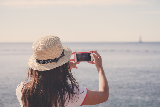 Young travelling woman in hat taking photos of summer sunny sea