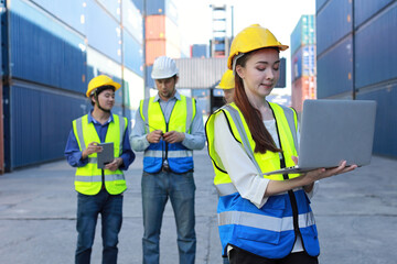 Group of multiethnic technician engineer in protective uniform standing and using computer, walkie talkie radio and tablet while.working together and controlling at container cargo site industrial