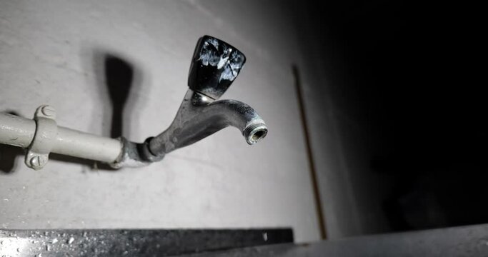 Old metal wall mounted faucet leaking water. Dark home cellar environment, close up, wide angle, slow motion, no people