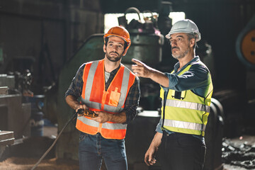 Senior male engineer training and explaining work to new employee wearing vest and safety jacket with hardhat helmet while pointing towards machine in factory and giving instructions - obrazy, fototapety, plakaty