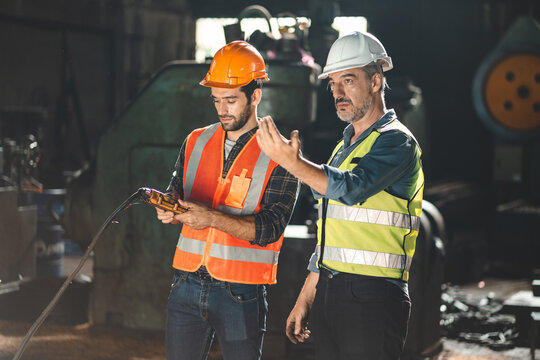 Senior male engineer training and explaining work to new employee wearing vest and safety jacket with hardhat helmet while pointing towards machine in factory and giving instructions
