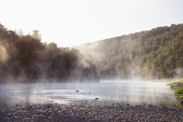 Silent beautiful morning summer landscape with cold blue mist on river with white fluffy haze on water and shine green forest on slopes in golden sunbeams. Majestic wild nature for travel.
