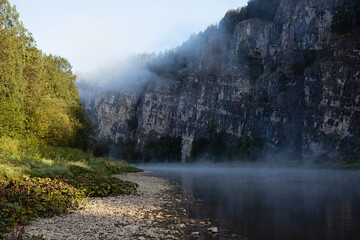 Majestic morning summer landscape in river canyon with rocky cliff in blue shadow and fog on river...