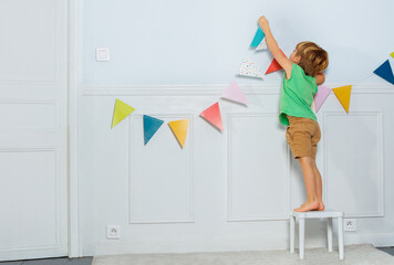 Young boy hangs birthday garland with flags on living room wall