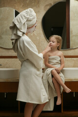 Portrait of mother with her little daughter wrapped in towels in the bathroom having fun during...
