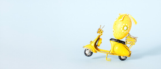 Small yellow figurine of motorbike with yellow and white Easter egg , on pastel blue background. Easter concept with copy space