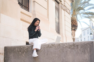 Young Asian girl studying with a laptop on the stairs of the university. She is drinking coffee. Concept of education.