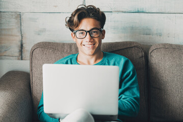 Portrait of cheerful young boy using laptop at home sitting on the sofa. One guy student using computer to have online class lessons with school comfortably sitting on couch. Smiling at the camera