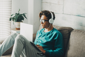 Fototapeta premium Relaxed young teenager using computer sitting on the sofa at home for play or study. Wearing headphones. One teenager boy listening music in indoor relax leisure activity alone. Afternoon time alone