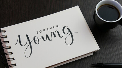 FOREVER YOUNG black lettering in notebook with cup of espresso and pens on black wooden desk - obrazy, fototapety, plakaty
