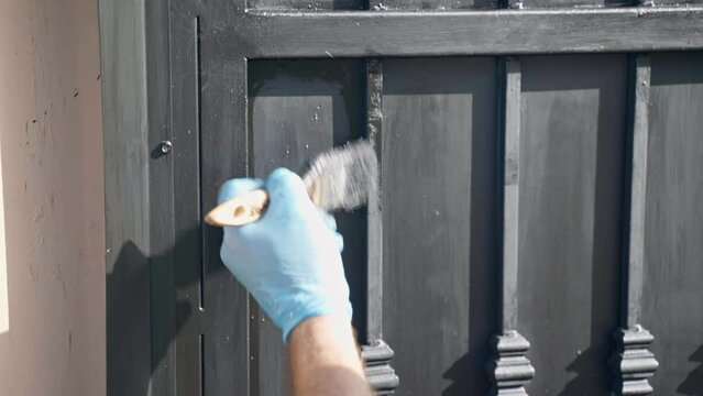 Workman painting restored metal gate with black colour using paintbrush, man in gloves coating steel surface with protecting paint, removing heavy corrosion and rust before final painting