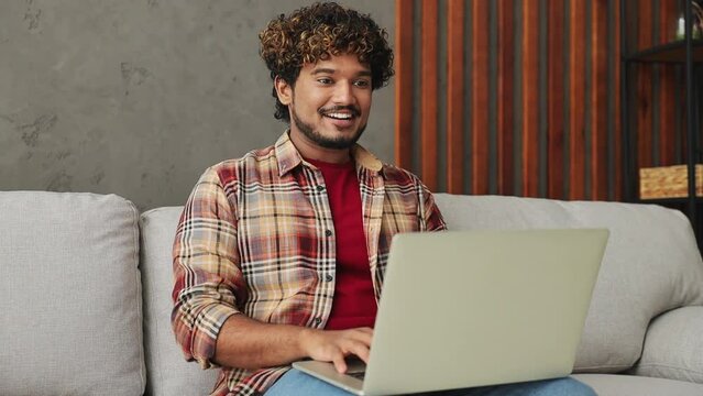 Smart IT user young Indian man wears casual clothes hold use work typing on laptop pc computer sits on grey sofa couch stay at home hotel flat rest relax spend free spare time in living room indoor