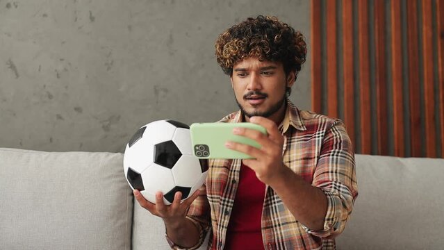 Young fun fan Indian man wears casual clothes cheer up support football sport team use mobile cell phone hold ball sits on sofa stay at home flat rest relax spend free spare time in living room indoor