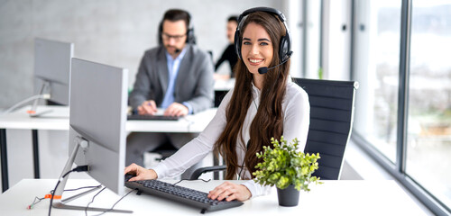 Online helpdesk worker in hands-free headset using computer in office. - obrazy, fototapety, plakaty