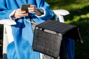 An unrecognizable woman with a mobile phone in her hands and a portable solar battery sits outdoors in the garden. Ecological alternative energy concept.
