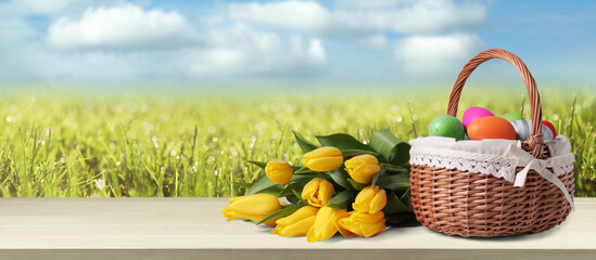 Wicker basket with festively decorated Easter eggs and tulips on wooden table on green meadow, space for text. Banner design