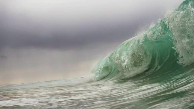 Large rough shorebreak wave crashes down on cloud filled stormy sky