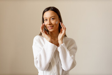 Attractive young woman in white bathrobe applies lotion or moisturizer to her face. Professional facial care after washing. Portrait of brunette girl on isolated background looking into the camera. - Powered by Adobe