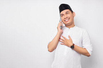 Friendly smiling young Asian Muslim man in Arabic clothes talking on mobile phone with hand his chest with grateful expression isolated on white background. People religious Islamic lifestyle concept