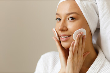 Young woman in white robe and towel on her head wipes the skin of her face with cotton pads. Cleansing and healthy body care after shower. Close-up of pretty girl on isolated background, copy space.