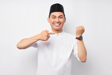 Smiling young Asian Muslim man in Arabic clothes pointing a finger at his wristwatch, indicate the time of the breaking fast isolated on white background. People religious Islamic lifestyle concept