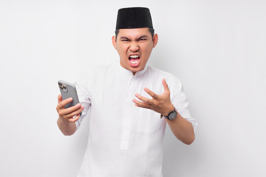 Upset young Asian Muslim man wearing Arabic clothes standing while using smartphone, screaming loudly when receiving bad news isolated on white background. People religious Islamic lifestyle concept
