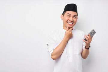 Smiling young Asian Muslim man holding mobile phone while looking side with touching chin and thinking about something isolated on white background. People religious Islamic lifestyle concept