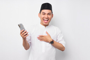 Smiling happy handsome young Asian Muslim man in Arabic clothes holding mobile phone and receiving good news isolated on white background. People religious Islamic lifestyle concept