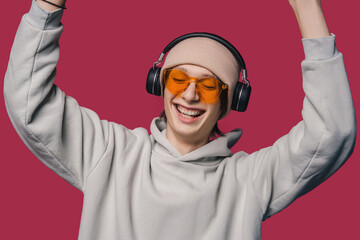 Photo of cool young guy dance in hat and sunglasses while listening his favorite music in headphones and smiling standing against pink background