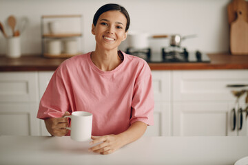 Cute young brunette woman in casual clothes looks straight into the camera. Portrait of smiling happy girl sitting at home in the kitchen at the dining table. Diet, fasting.