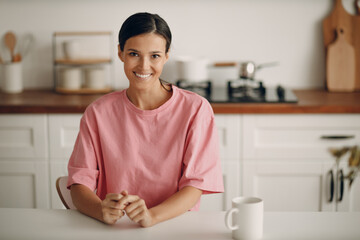 Fototapeta premium Spanish young brunette woman in casual clothes looks straight into the camera. Portrait of smiling happy girl sitting at home in the kitchen at the dining table. Diet, fasting.