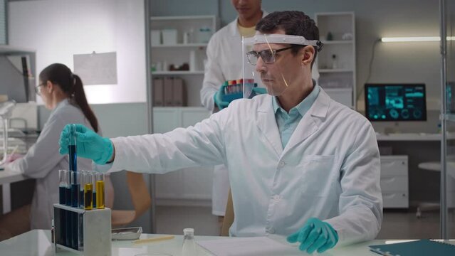 Waist up shot of professional Caucasian male microbiologist in protective face shield and white lab coat examining culture of liquids in glass test tubes, working in scientific lab with assistant