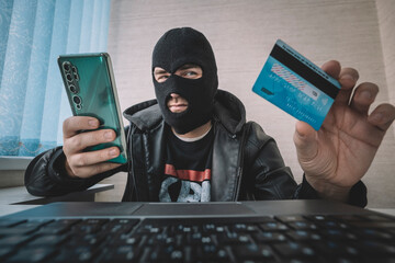 Male hacker in a robber mask uses phone, credit card and laptop in some fraudulent scheme. Cyber...