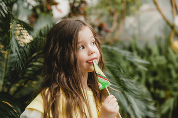 Cute little girl eating a watermelon shaped lollipop Child with lollipops in the botanical garden.