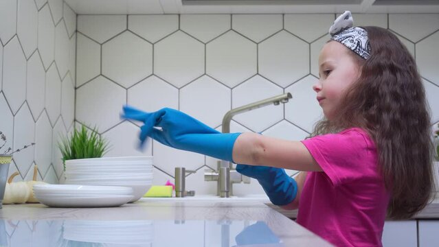 Side view of little preteen girl with loose long dark hair wearing pink T-shirt, putting on big blue rubber gloves for washing dishes near stack of clean white plates in kitchen. Housework, cleaning.