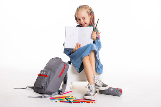 Happy preschooler kid ready for school. Happy family concept kid in good mood sitting on white floor in studio isolated holding pencils pens notebook in hands showing at camera isolated.