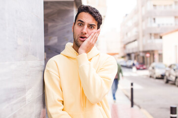 young hispanic man feeling shocked and astonished holding face to hand in disbelief with mouth wide open